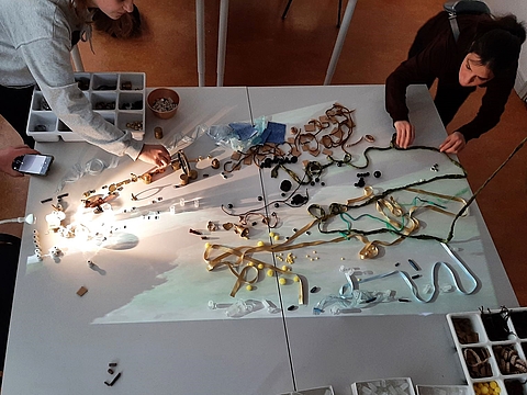 Zwei erwachsene Personen haben auf einem großen, weißen Tisch Muster aus vielen Alltagsgegenständen und vermeintlichem "Abfall" gelegt. Mit einer Klemmlampe werden die Materialien ausgeleuchtet und mit einem Smartphone als Video aufgenommen.